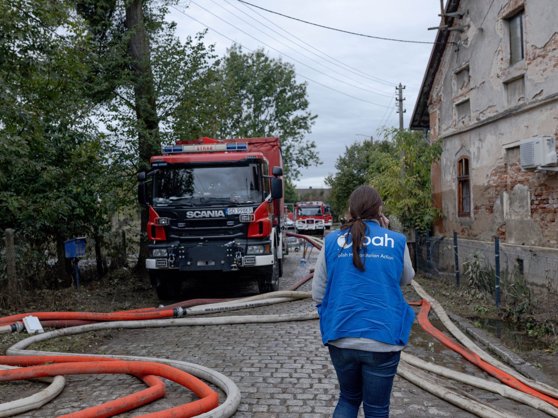 Pracowniczka PAH w niebieskiej kamizelce, dalej wóz strażacki, węże do pompowania wody leżą na ziemi obok wozu.